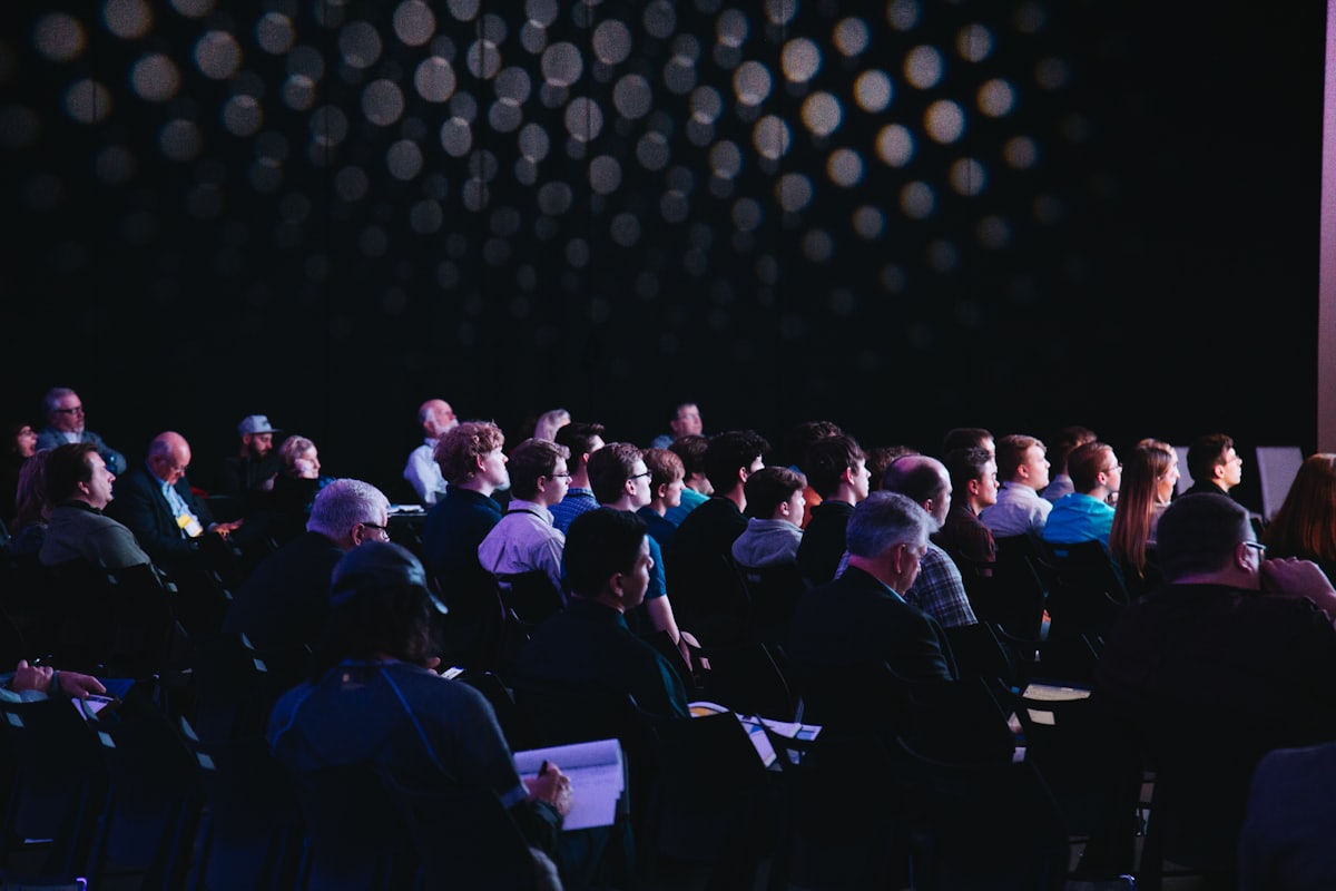 An audience seated for an evening talk in a darkened room lit by a wall of warm bulbs