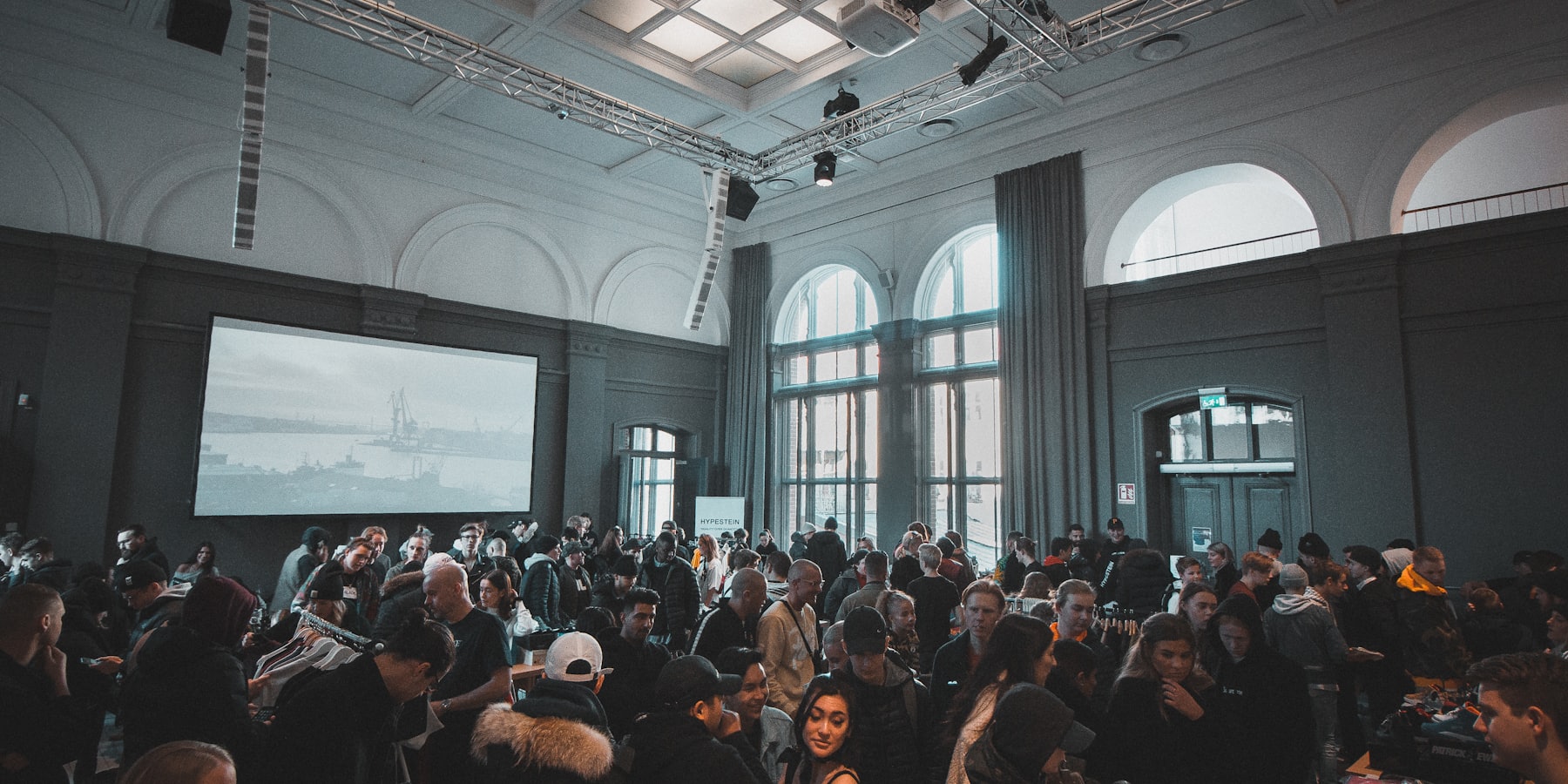 A full room at an evening event in The Gallery, lit by the screen and the arched windows